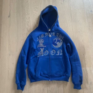 Blå hoodie från Aim for the Moon - Säljer en blå hoodie från Aim for the Moon med dragkedja och tryck på framsidan. Hoodien har en klassisk passform med långa ärmar och en bekväm huva. Perfekt för en avslappnad stil.