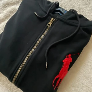 Svart hoodie från Polo Ralph Lauren - Säljer en svart hoodie från Polo Ralph Lauren med dragkedja och en stor röd logga på bröstet. Tröjan har långa ärmar och en huva med dragsko. Tillverkad i 100% bomull för en bekväm passform. Perfekt för en avslappnad stil.