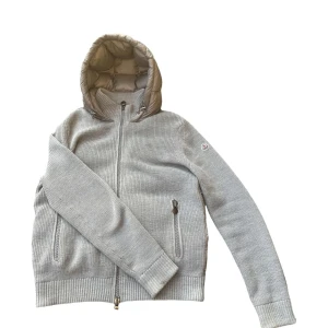 Grå jacka från Moncler - Säljer en snygg grå jacka från Moncler med stickade ärmar och en quiltad rygg. Jackan har en huva och dragkedja framtill. Perfekt för kyligare dagar.