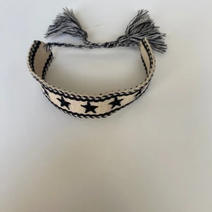 Beige och svart armband med stjärnor - Snyggt svart och vitt armband med svarta stjärnor och flätade tofsar. Perfekt accessoar för att ge din outfit en unik touch. Justerbar passform med knytning.