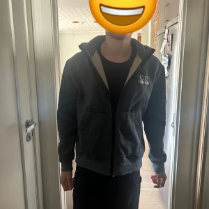  C.P. Company zip - Säljer en grå huvtröja från C.P. Company med dragkedja och coola glasögondetaljer på huvan. Perfekt för en avslappnad stil. Tröjan har långa ärmar och en normal passform.