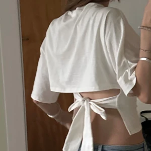 Vit croppad topp med knyt i ryggen - Säljer en vit croppad topp med korta ärmar och rund hals. Toppen har en öppen rygg med breda band som knyts i en rosett bak, vilket ger en snygg och trendig look. 