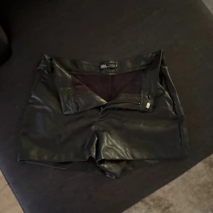 Svarta skinnshorts från Zara - Snygga svarta shorts i skinnimitation från Zara. De har en klassisk passform med dragkedja och knapp framtill. Perfekta för dig som vill ha en edgy och trendig look. 
