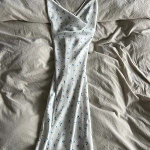 Vit blommig långklänning från NA-KD - Superfin långklänning i vitt med små blå blommor från NA-KD. Klänningen har draperande ringning, smickrande omlott-detalj och snörning i ryggen. Perfekt för sommarens alla tillfällen och ger en romantisk vibe.