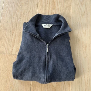 Mörkgrå kofta från Gran Sasso - Mörkgrå kofta från Gran Sasso med hel dragkedja framtill och ribbade muddar. Tillverkad i mjukt material som känns skönt mot huden. Perfekt för lager-på-lager och har en stilren, enkel design med hög krage.