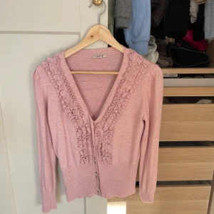 Rosa kofta med volang från Selected Femme - Supersöt ljusrosa kofta från Selected Femme med volangdetaljer längs v-ringningen och knappar framtill. Långärmad och ribbad vid ärmslut och midja. Perfekt för dig som gillar en feminin och trendig look.