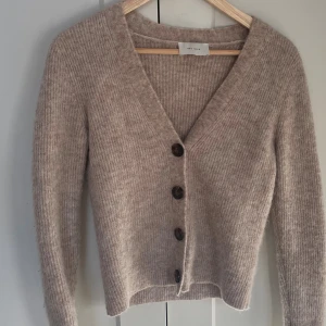 Beige kofta från Neo Noir - Supermjuk och ribbstickad beige kofta från Neo Noir med v-ringning och stora mörka knappar framtill. Långärmad och i en lite kortare modell som är perfekt att slänga över en topp. Gjord i ett fluffigt och mysigt material.