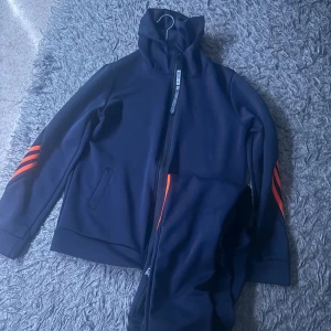 Mörkblå träningsjacka med orange detaljer - Mörkblå träningsjacka från New8 i storlek M. Jackan har dragkedja framtill, hög krage och orange ränder på ärmarna. Tillverkad i mjukt syntetmaterial och har två fickor på sidorna. Perfekt för dig som gillar sportig stil.