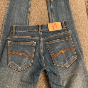 Blå jeans från Nudie Jeans - Säljer ett par klassiska blå jeans från Nudie Jeans med snygga orange sömmar och vågdetalj på bakfickorna. Jeansen har en rak passform och är tillverkade i slitstarkt denimtyg. Perfekta för dig som gillar stilrena och tidlösa jeans.