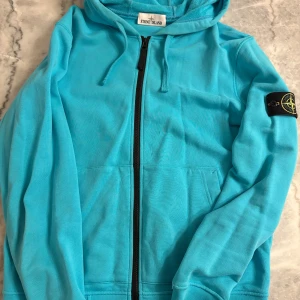 Turkos hoodie från Stone Island - Säljer en turkos hoodie från Stone Island med dragkedja och huva. Tröjan har klassisk logga på ärmen, känguruficka framtill och snörning i huvan. Perfekt för dig som gillar streetwear och vill sticka ut med färg.