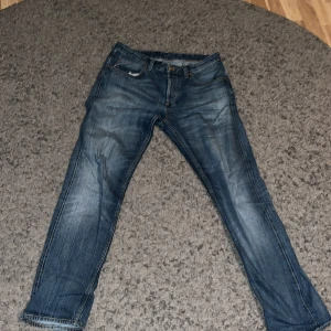 Blå jeans från Gant med raka ben - Säljer ett par klassiska blå jeans från Gant med snygga slitningar och raka ben. Jeansen har fem fickor, detaljer med kontrastsömmar och en läderpatch med Gant-logga bak i midjan. Perfekt för dig som gillar en tidlös och avslappnad stil.