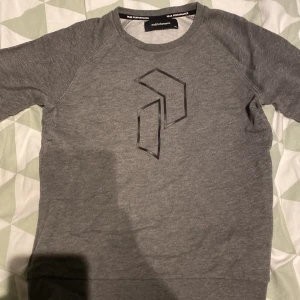 Grå sweatshirt från Peak Performance - Snygg grå sweatshirt från Peak Performance med deras ikoniska logga tryckt på bröstet. Tröjan har rund halsringning och långa ärmar med muddar. Perfekt för en chill och sportig stil.