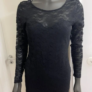 Svart spetsklänning med lång ärm - Svart kort klänning i figurnära modell med långa ärmar och helspets över hela plagget. Klänningen har rund halsringning och transparenta spetsdetaljer på ärmar och axlar. Perfekt för dig som gillar en elegant och stilren look.