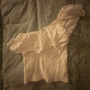 Vit offshoulder topp med volang - Säljer en vit offshoulder topp med bred volang och broderade blommor längs kanten. Toppen är ärmlös och har en asymmetrisk design med en axel bar. Perfekt för dig som gillar romantiska detaljer och ljusa färger.