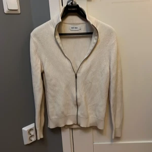 Vit ribbad kofta från East West - Säljer en stilren vit ribbad kofta från East West i storlek S. Koftan har hel dragkedja framtill, hög krage och är långärmad. Perfekt att slänga på sig när det är lite kyligare ute. Materialet är stickat och känns mjukt mot huden.