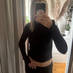 Svart långärmad topp - Säljer en svart långärmad topp med tight passform. Är lite högre i halsen och en slot vid ärmens slut. Toppens material känns mjukt och följsamt mot kroppen.