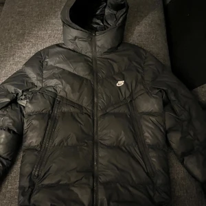 Svart pufferjacka Nike Storm-FIT S - Svart pufferjacka från Nike med Storm-FIT-teknologi. Jackan har huva, två stora dragkedjefickor framtill och klassisk Nike-logga på bröstet. Perfekt för kalla dagar med sin vadderade design och sportiga look.Mäket på jackan har gått sönder lite. Nypris 2500kr