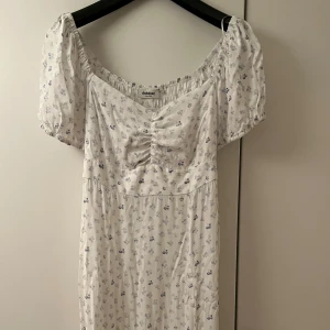 Vit blommig långklänning från Dobber - Romantisk långklänning från Dobber i vit med små blå blommor, puffärmar och smockad rygg. Klänningen har v-ringning med rynkdetalj framtill och är tillverkad i ett mjukt, lätt tyg. Klänningen har en slits längt ner på ena sidan. Perfekt för sommarens alla tillfällen. Storlek S