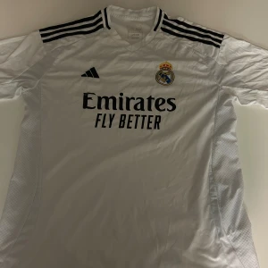 Real Madrid matchtröja Adidas 2XL - Vit Real Madrid fotbollströja från Adidas med svarta ränder på axlarna och klubbmärke broderat på bröstet. Tryck med 'Emirates Fly Better' på framsidan. Klassisk V-ringning och tillverkad i lätt, ventilerande material.
