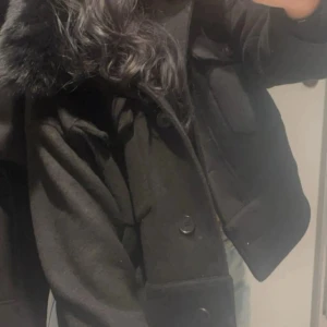 Svart jacka med fuskpäls-krage - Snygg svart jacka med stor fuskpäls-krage och knappar framtill. Jackan har två bröstfickor med lock och en croppad passform. Perfekt för dig som vill ha en stilren och trendig look under kyliga dagar.