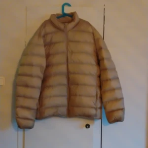 Beige pufferjacka med dragkedja - Säljer en beige pufferjacka med hög krage och horisontella sömmar. Jackan har en enkel design med dragkedja framtill och långa ärmar. Perfekt för kalla dagar när du vill hålla värmen och ändå se stilren ut.