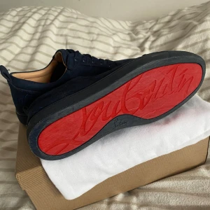 Mörkblå mocka sneakers Louboutin - Snygga mörkblå sneakers från Christian Louboutin i lyxig mocka med klassisk röd sula och diskret logga på hälen. Skorna har rund tå, platt sula och snörning. Kommer med originalkartong och dustbag. Såklart äkta. Perfekta för dig som vill sticka ut med stil. 