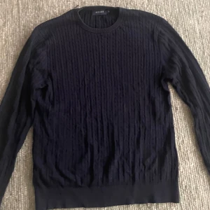 Mörkblå kabelstickad tröja Riley - Säljer en mörkblå kabelstickad tröja från Riley i storlek M. Tröjan har rund halsringning, ribbade muddar och lång ärm. Perfekt för dig som gillar klassisk och stilren look med lite struktur i materialet. NYPRIS: 900kr