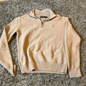 Beige stickad tröja Polo Ralph Lauren - Beige stickad tröja från Polo Ralph Lauren med half zip och klassisk broderad logga på bröstet. Tröjan har lång ärm, ribbade muddar och hög krage. Perfekt för lager-på-lager och snygg till jeans eller chinos.