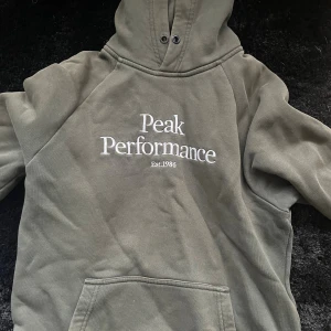 Grön hoodie från Peak Performance - Säljer en grön hoodie från Peak Performance med stor vit logga broderad på bröstet. Klassisk känguruficka framtill och huva med snörning. Perfekt för chill dagar eller när du vill ha en avslappnad stil.