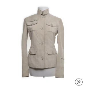 Beige mockajacka med ståkrage - Snygg beige jacka i mocka med ståkrage och bälte i halsen. Jackan har fyra fickor framtill, samt dekorativ knappdetalj i ryggen. Figurnära passform och långa ärmar. Perfekt för dig som gillar stilren och clean look. (Det finns tecken på användning därav de billiga priset)