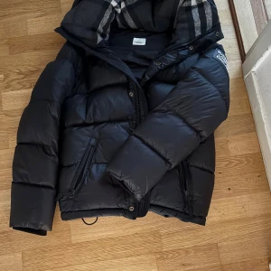 Svart pufferjacka från Burberry - Säljer en svart pufferjacka från Burberry med klassiskt rutigt foder i huvan och tydlig logga på ärmen. Jackan har avtagbara ärmar så den kan bli en väst top skick 9/10 hojta till vid frågor pris ej hugget i sten. äkta