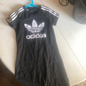 Svart Adidas klänning med logga - Svart kortklänning från Adidas med stor vit trefoil-logga och text framtill. Klassiska vita ränder på axlarna och korta ärmar. Tillverkad i mjuk bomull, perfekt för en sportig och chill stil. Rund hals och normal passform.