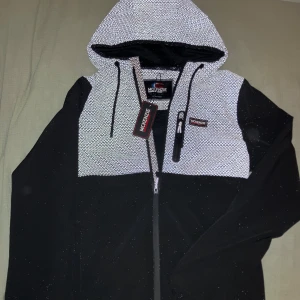 Svart vindjacka från McKenzie med reflex - Svart och vit vindjacka från McKenzie med reflekterande mönster upptill och huva med snörning. Jackan har hel dragkedja, bröstficka med dragkedja och sportig look. Perfekt för dig som gillar streetwear och vill sticka Utes en riktigt fet slim fit jacka