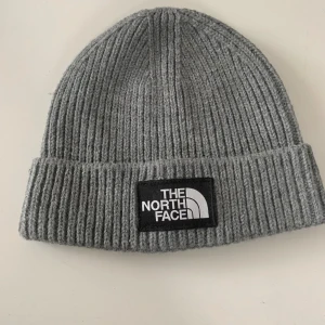 The North Face Mössa - Säljer en grå ribbstickad mössa från The North Face. Klassisk modell med uppvikt kant och tydlig svart logotyp-patch framtill. Tillverkad i mjuk och stretchig syntetblandning som sitter skönt på huvudet. Perfekt för kalla dagar.