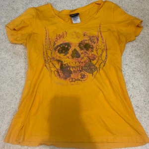 Gul Harley-Davidson t-shirt med tryck - Gul t-shirt från Harley-Davidson med coolt döskalletryck och blommor framtill, samt lila tribal-tryck och logga på ryggen. Skön passform och tillverkad i mjuk bomull. Storlek L men passar även M