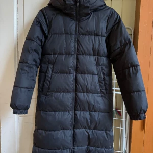 Svart lång pufferjacka från Everest - Lång svart pufferjacka från Everest med huva och dragkedja framtill. Jackan har quiltad design, två snedställda fickor och elastiska muddar vid ärmsluten. Perfekt för kalla dagar och riktigt skön att bära.