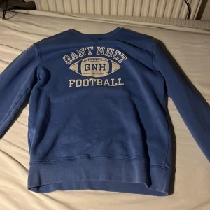 Blå sweatshirt från Gant med tryck - Blå sweatshirt från Gant med stort vitt tryck på bröstet där det står 'GANT NHCT FOOTBALL' och en amerikansk fotboll. Tröjan har rund halsringning och långa ärmar. Perfekt för en sportig och avslappnad stil.