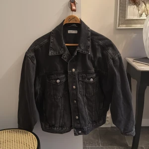 Svart jeansjacka från Gina tricot - Säljer en svart jeansjacka från Perfect Jeans i storlek M. Jackan har klassisk krage, silverfärgade knappar framtill och två bröstfickor med lock. Snygg, lite loose passform och tillverkad i robust denim. Perfekt för dig som gillar en tidlös streetstil.