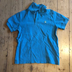 Blå pikétröja från Benetton - Säljer en blå pikétröja från Benetton med klassisk krage och knappar framtill. Tröjan har korta ärmar och en liten broderad logga på bröstet. Perfekt för en avslappnad och stilren look.