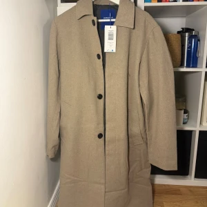 Beige rock från Jack & Jones - Stilren beige rock från Jack & Jones med klassisk krage och svarta knappar framtill. Jackan har raka linjer, två sidfickor och är helfodrad. Perfekt för dig som gillar en clean och tidlös look. Stor i storlek