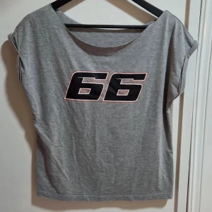 Grå loose topp med tryck 66, M - Säljer en grå loose topp med stort svart tryck '66' på bröstet, omringat av en tunn röd linje. Toppen har bred halsringning och är kortärmad med lätt upprullade ärmslut. Mjuk och skön bomullskänsla, perfekt för chill dagar.