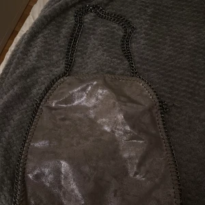 Grå axelväska med kedjedetaljer - Snygg grå axelväska med glansig yta och coola kedjehandtag. Väskan har dragkedja upptill och dekorativa kedjor längs kanterna som ger en edgy vibe. Perfekt storlek för att få plats med allt du behöver till vardagen.