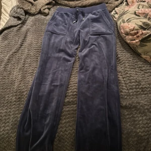 Blå velourbyxor från Juicy Couture - Mörkblå mjukisbyxor i velour från Juicy Couture med bred resår och snörning i midjan. Byxorna har raka ben, stora fickor fram och broderad JC-logga på ena fickan. Metalländar på snörena med Juicy Couture-gravyr för extra detalj.