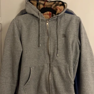 Grå hoodie från Burberry med dragkedja - Säljer en grå hoodie från Burberry med klassiskt rutigt foder i huvan. Tröjan har dragkedja framtill, snörning i huvan och två stora fickor. Perfekt för dig som gillar stilren streetwear med en touch av lyx, Skicket är 6/10 därav priset, den har sina skavanker som man får skriva privat för att få bilder på, Priset är inte hugget i sten