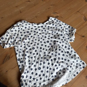 Vit t-shirt med blå prickar - Vit t-shirt med mörkblå prickigt mönster. Klassisk rund halsringning och korta ärmar. Skön och luftig passform, perfekt för varma dagar. T-shirten är i mjukt bomullsmaterial och har en avslappnad vibe.