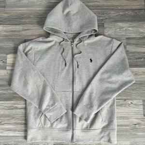 Ralph lauren zip - Snygg grå hoodie från Polo Ralph Lauren med dragkedja framtill, huva med snörning och klassisk broderad logga på bröstet. Tillverkad i mjuk bomullsmix och har två fickor framtill. Perfekt för en chill och stilren look.