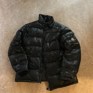 Svart pufferjacka från Fashion - Svart pufferjacka med glansig finish och hög krage. Jackan har en patch med Fashion-logga på ärmen, dragkedja framtill och praktiska fickor. Perfekt för kalla dagar och ger en cool vibe till din outfit.