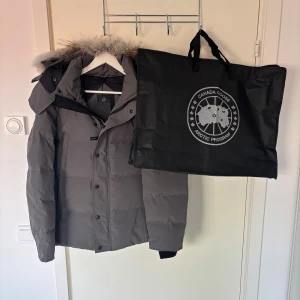 Grå Canada Goose wyndham - Säljer en grå Canada Goose Wyndham Parka i storlek M. Jackan har en klassisk parkamodell med huva, svarta knappar och dragkedja.  Dustbag ingår vid köp