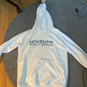 Vit hoodie från McKenzie med tryck - Vit hoodie från McKenzie med blått och svart tryck på bröstet där det står 'McKenzie Original Sportswear'. Tröjan har huva, lång ärm och stor magficka framtill. Perfekt för chill dagar och streetstyle. Materialet känns mjukt och skönt mot huden.