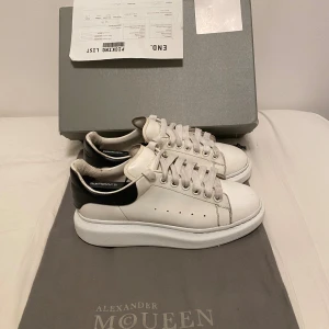 Alexander McQueen vita sneakers - Säljer ett par klassiska Alexander McQueen sneakers i vitt skinn med svart häl och tjock vit plattformssula. Skorna har rund tå, snörning och diskreta hål på sidorna för ventilation. Perfekta för dig som gillar stilrena och trendiga sneakers.                                                           Passar storlek 42.                                                  Allt utom box ingår kan även ta byten 